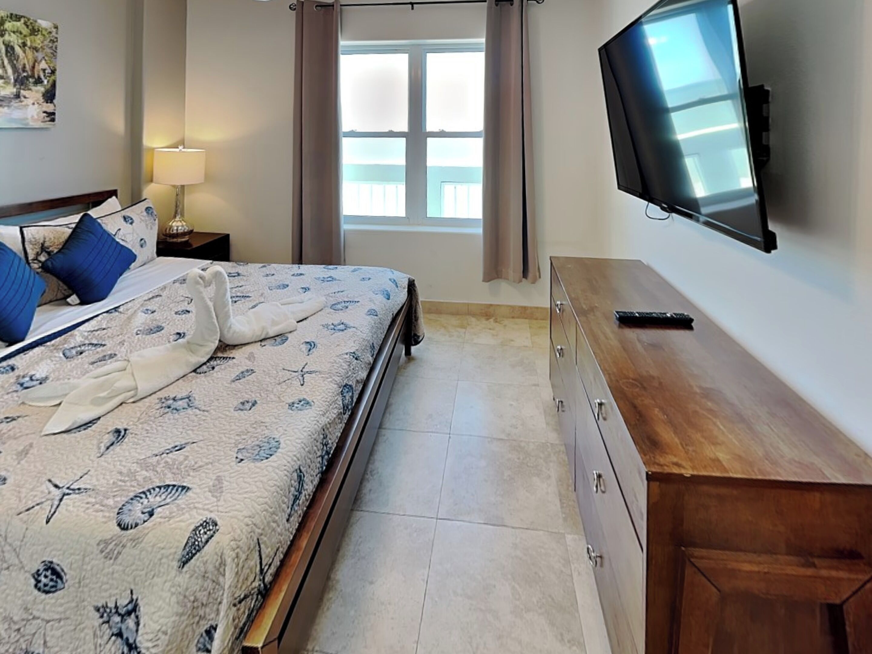 Une chambre à coucher comprenant un lit, un téléviseur, une commode en bois, une table de chevet, une lampe et une fenêtre avec des rideaux.