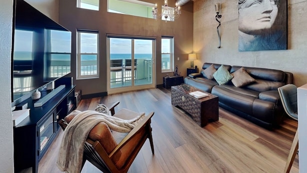 A spacious living area with a large TV, a sofa, a coffee table, and a view of the ocean through large windows. - Ocean Grove PH 1101 - The Sky Gallery Penthouse (Galveston)