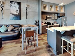 A modern kitchen with a central island, a dining area with a table and chairs, and a living room with a sofa and a large painting. - Ocean Grove PH 1101 - The Sky Gallery Penthouse (Galveston)