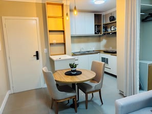 A compact kitchen with white cabinets, a dining table with two chairs, a potted plant, and a hanging light fixture. - Ravello Residencial – Comfort and tranquility in Foz do Iguaçu (Foz do Iguaçu)