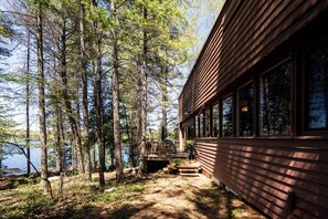 A cabin with a deck overlooking a lake, surrounded by tall trees. - Cozy Lakefront Cottage | Firepit, Dock & Lake Access (Lake of Bays)