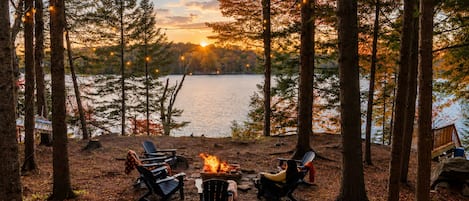Un campamento con un hoyo para asar, sillas y vistas a un lago rodeado de árboles.