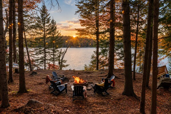 Un terrain de camping équipé d’un foyer, de chaises et offrant une vue sur un lac entouré d’arbres.