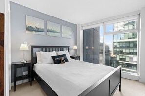 Ein modernes Schlafzimmer mit einem großen Bett, zwei Nachttischen, einem Beistelltisch, Blick auf eine Stadtlandschaft und einem großen Fenster.