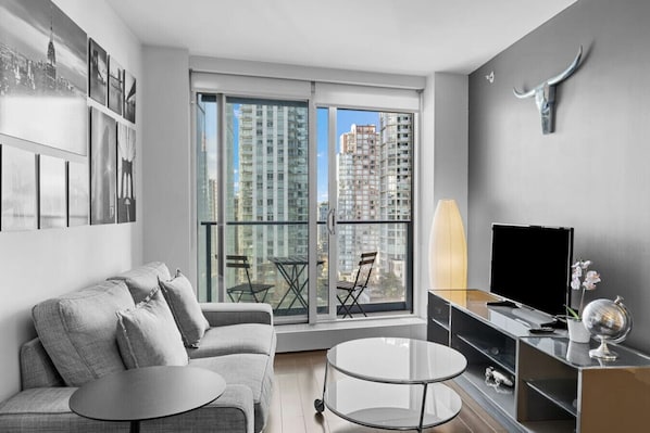 Ein modernes Wohnzimmer mit einem grauen Sofa, einem Glastisch, einem Flachbildfernseher und einem großen Fenster mit Blick auf die Stadt.