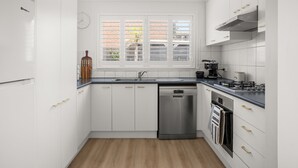 Een moderne keuken met witte kastjes, een vaatwasser, een fornuis en een raam met luxaflex.