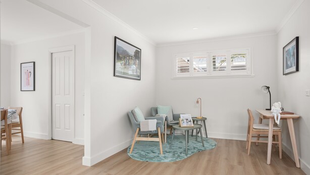 Een moderne woonkamer met een bank, stoelen, een klein tafeltje en een vloerkleed. Er hangen fotolijsten aan de muren en een deur leidt naar een andere kamer.