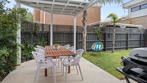 Een overdekt terras met een houten tafel en witte stoelen, een barbecue en een bakstenen gebouw op de achtergrond.