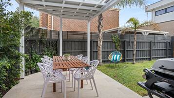 A covered patio with a wooden table and white chairs, a grill, and a brick building in the background.
