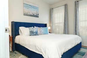 Une chambre à coucher avec une tête de lit bleue, du linge de lit blanc et un tableau représentant des fleurs accroché au mur.