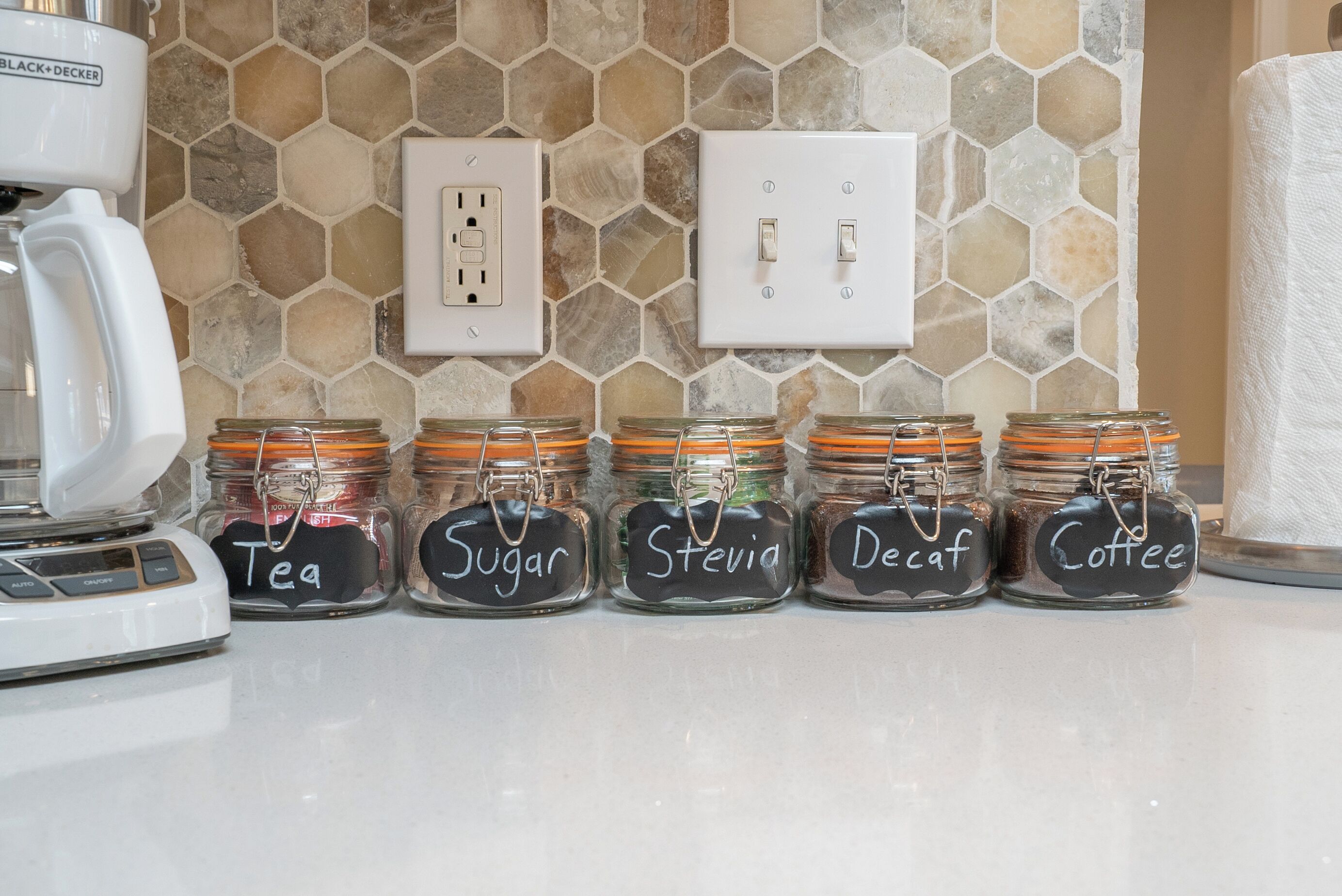 Un piano di lavoro in cucina con cinque barattoli di vetro etichettati con diversi tipi di tè, zucchero, stevia, decaffeinato e caffè.