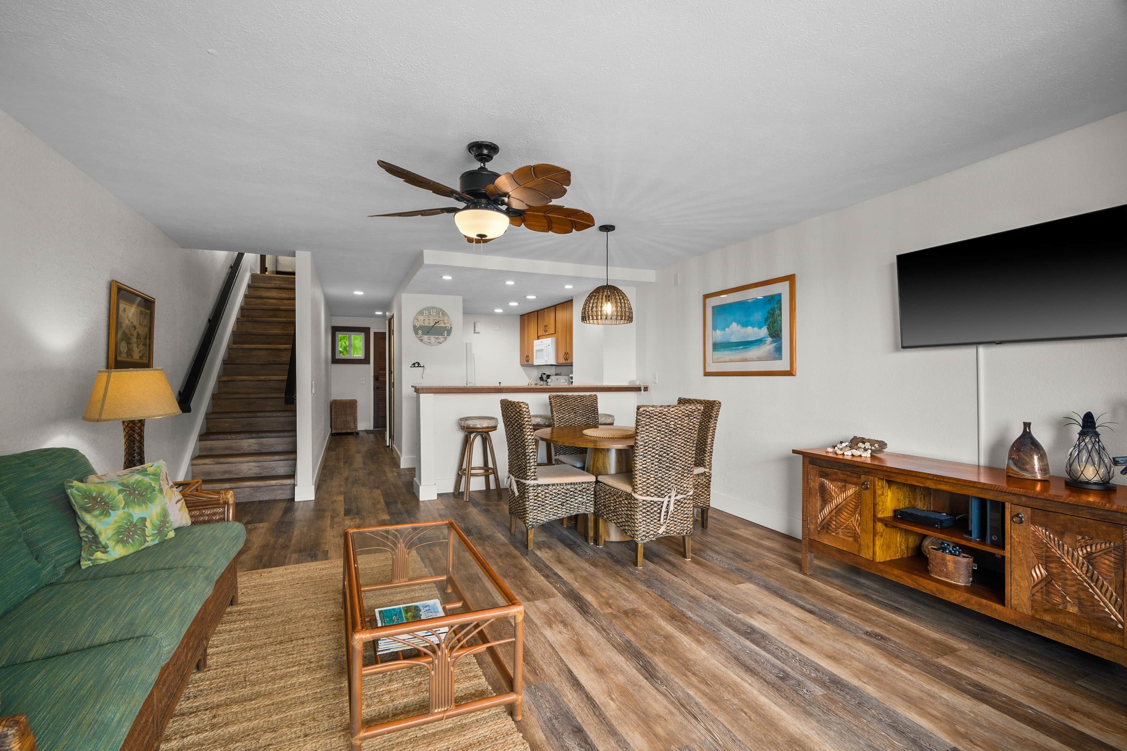 A living room with a sofa, coffee table, dining table, chairs, a ceiling fan, a TV, and a staircase leading to a kitchen.