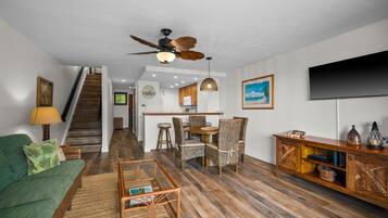 A living room with a sofa, coffee table, dining table, chairs, a ceiling fan, a TV, and a staircase leading to a kitchen.