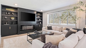 A modern living room with a flat-screen TV, dark wood shelving, a glass coffee table, and a large window with a view. - Private Pool, Infared Sauna, Full Gym, Hot Tub, Sleeps 16 (Grandville)