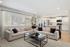 A modern living room with a sofa set, a coffee table, and a kitchen in the background. - Private Pool, Infared Sauna, Full Gym, Hot Tub, Sleeps 16 (Grandville)