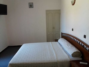 A single bed with a wooden headboard, a white mattress, and a pillow. There is a closed door in the background and a wall-mounted light fixture.