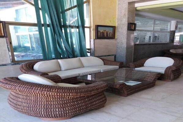 Un conjunto de sofá de mimbre con cojines, una mesa de centro de vidrio y vistas a un edificio a través de amplios ventanales.