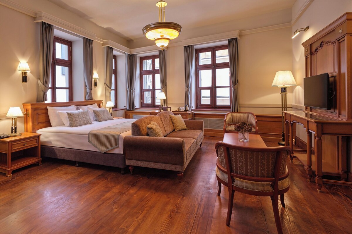 A hotel room with a large bed, a sofa, a TV, and a desk with a lamp.