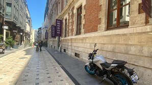 Asfaltoitu kävelytie, jonka varrella on pysäköity moottoripyörä, kylteillä varustettuja rakennuksia ja kirkas taivas.