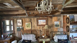 Une pièce rustique aux murs en bois, avec deux lits recouverts de draps à carreaux, un lustre et des tableaux encadrés.