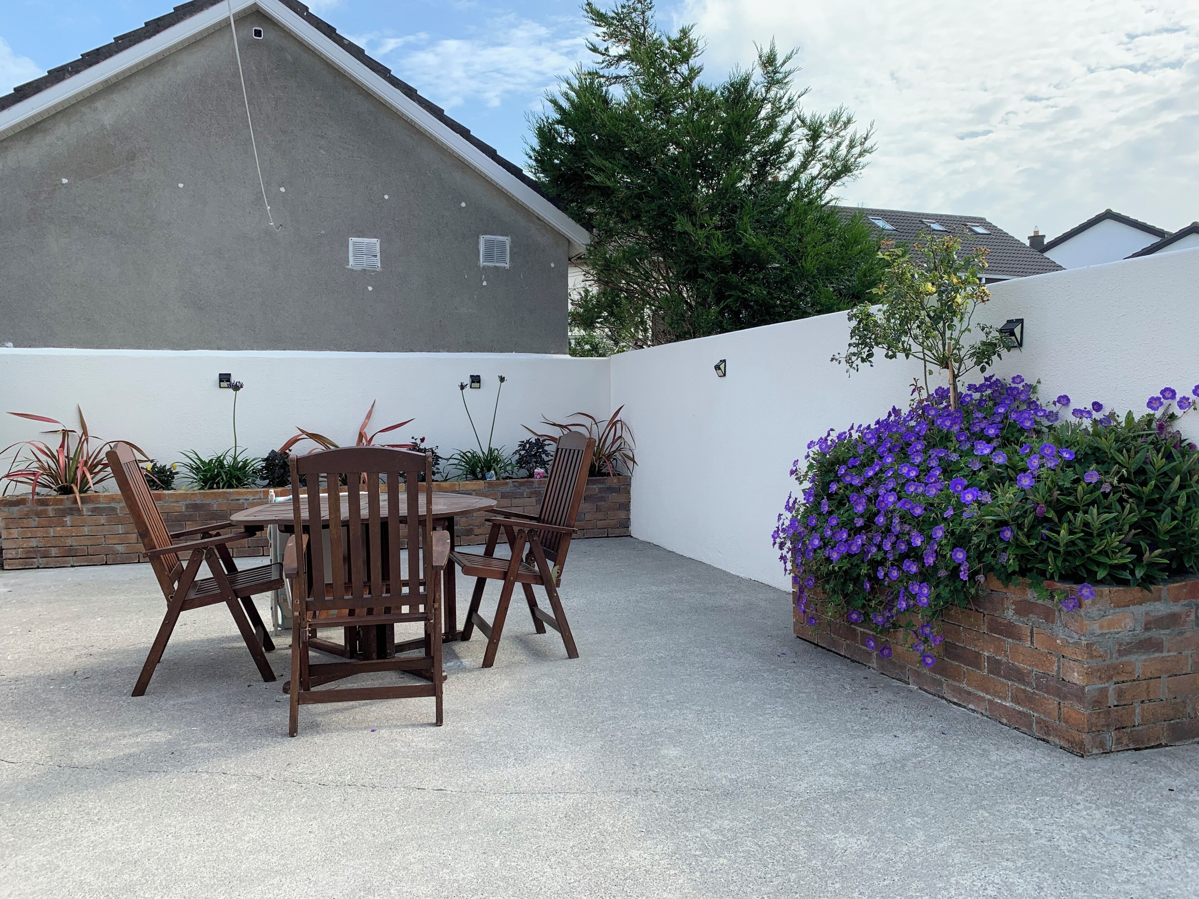 Eine Terrasse mit Tisch und Stühlen, ein Ziegelkübel mit violetten Blumen und eine weiße Wand.