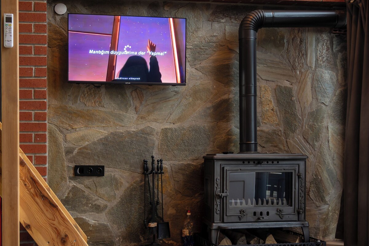 Una pared de piedra con un televisor montado encima, una chimenea con su correspondiente chimada, y una escalera de madera.