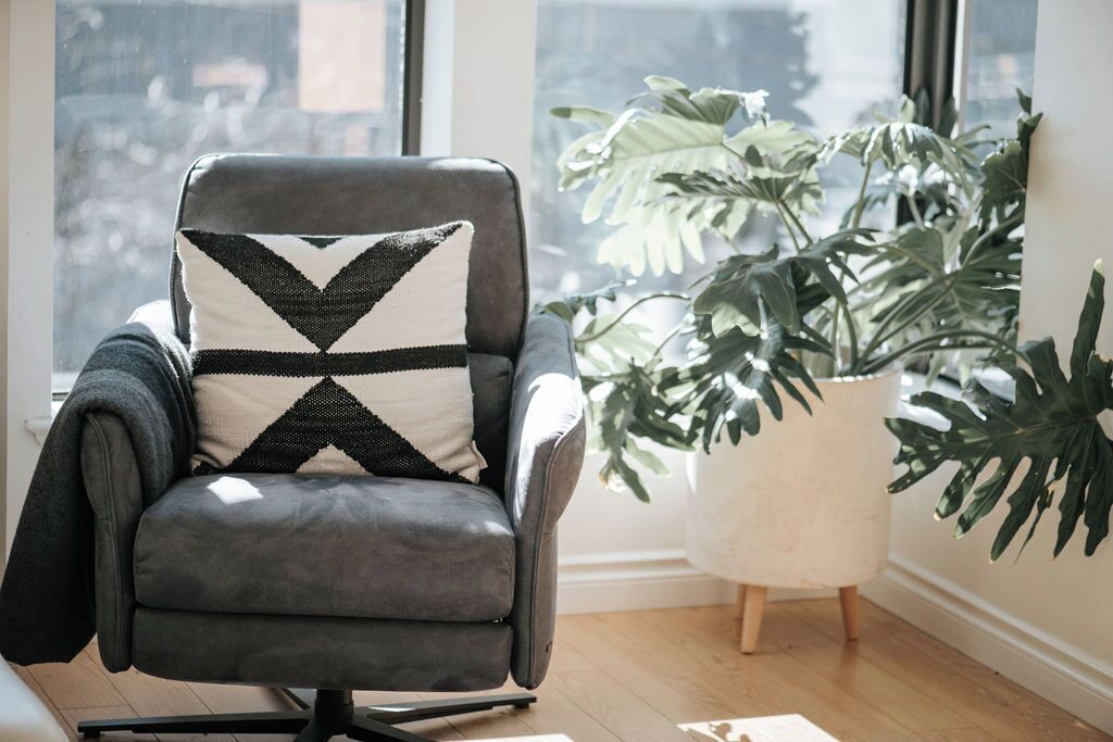 A grey armchair with a black and white geometric pillow, a large potted plant, and a window with a view of buildings.