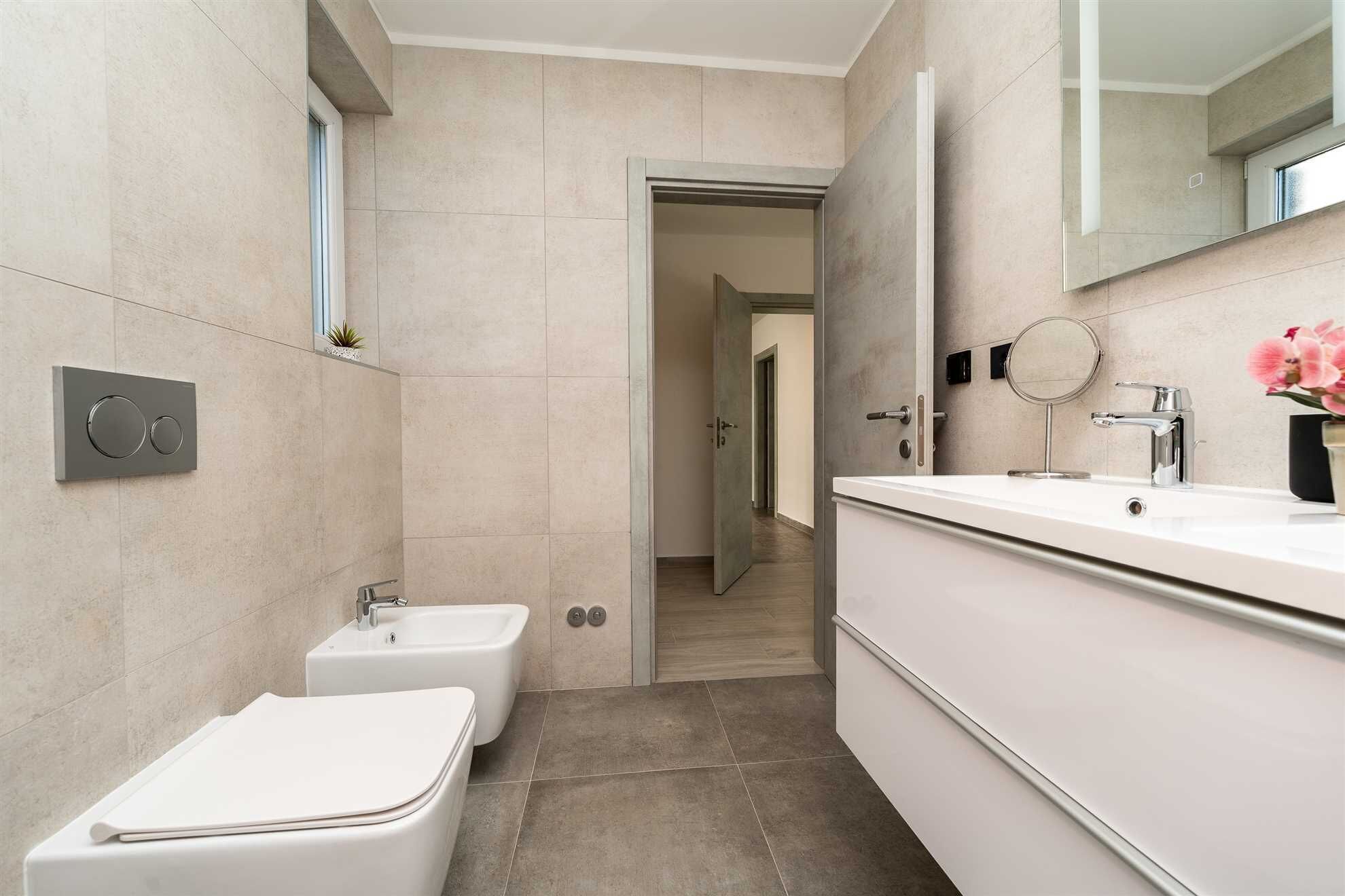 Een moderne badkamer met toilet, bidet en wastafel. Er is een spiegel, een kleine plant en een deur naar een andere ruimte.