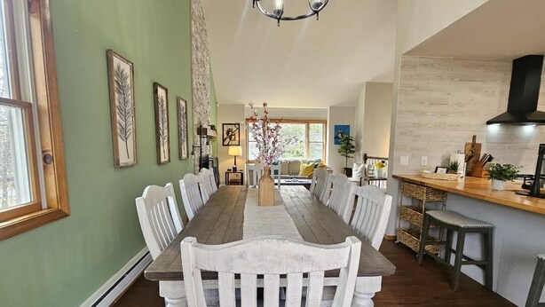 A dining area with a long wooden table, white chairs, a chandelier, and a kitchen area with a counter and stools. - Arrowhead Community Hot Tub Firepit Game Rm Movie Rm Fireplace Walk 2 Pool, Lake (Pocono Lake)