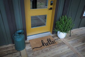 A yellow door with a glass panel, a green trash can, a potted plant, and a doormat with the word 'hello'.