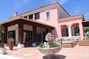 Eine rosa Villa mit überdachter Terrasse, Außenmöbeln und einem großen Pflanzgefäß mit Pflanzen.