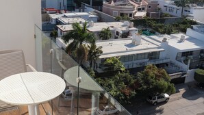 Ein Balkon mit Glasgeländer, einem runden Tisch und einem Stuhl, mit Blick auf einen Parkplatz mit mehreren Autos und Gebäuden im Hintergrund.