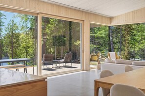 A modern interior with large glass doors leading to an outdoor deck, wooden furniture, and a view of a forest. - The Lost Lake Residence (Wentworth-Nord)