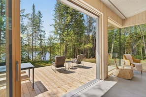 A modern cabin with a deck, a table, and chairs, overlooking a forest and a lake. - The Lost Lake Residence (Wentworth-Nord)