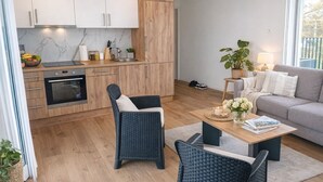 A modern kitchen with white cabinets, a built-in oven, and a marble backsplash. There is a dining area with wicker chairs and a wooden coffee table. A grey sofa and a potted plant are visible in the living room area. - Modern Studio Zurich Center (Zürich)