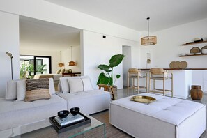Una sala de estar moderna con sofá, reposapiés y mesa de centro, un comedor con sillas y una cocina con estanterías.