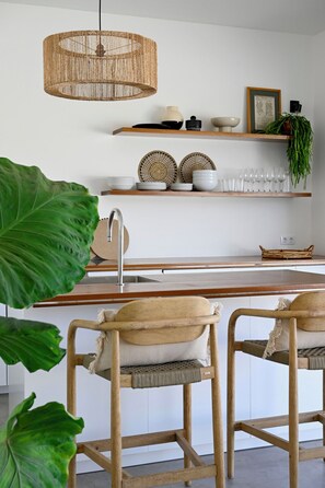 A modern kitchen with a central island, wooden chairs, a hanging light fixture, and a potted plant.