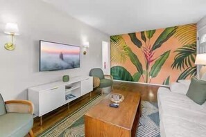 A modern living room with a flat-screen TV, a sofa, a coffee table, and a wall with a tropical mural.