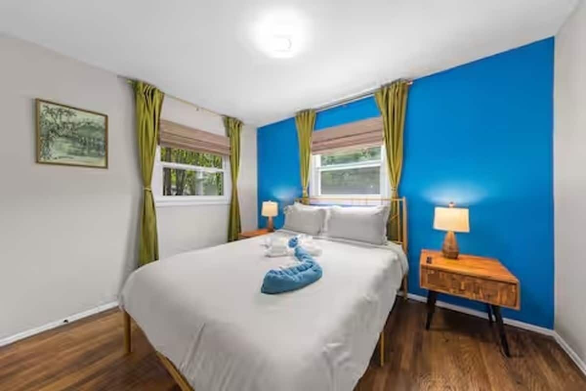 Una habitación de hotel con una cama, dos mesitas de noche, una ventana con cortinas y un cuadro en la pared.