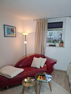 Een rode leren bank met een witte plaid, een glazen salontafel met boeken en een tijdschrift, een vloerlamp, een raam met uitzicht op bomen en een lijst met een schilderij aan de muur.