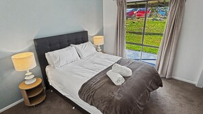 A neatly made bed with white linens and a dark gray throw, two bedside lamps, a small wooden nightstand, and a view of a red car and greenery outside through a window with curtains.