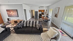 A modern living room with a dark sofa, a wooden coffee table, and a fluffy chair. There is a dining area with a wooden table and chairs. The room has white walls and a large window.