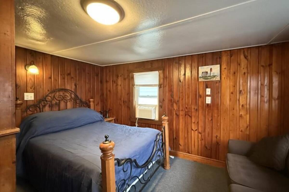 A room with wooden paneling, a bed with a headboard, a window, and a wall-mounted picture.
