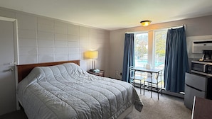 Quarto de hotel com cama, escrivaninha, cadeira, luminária, janela com cortinas e cozinha compacta.