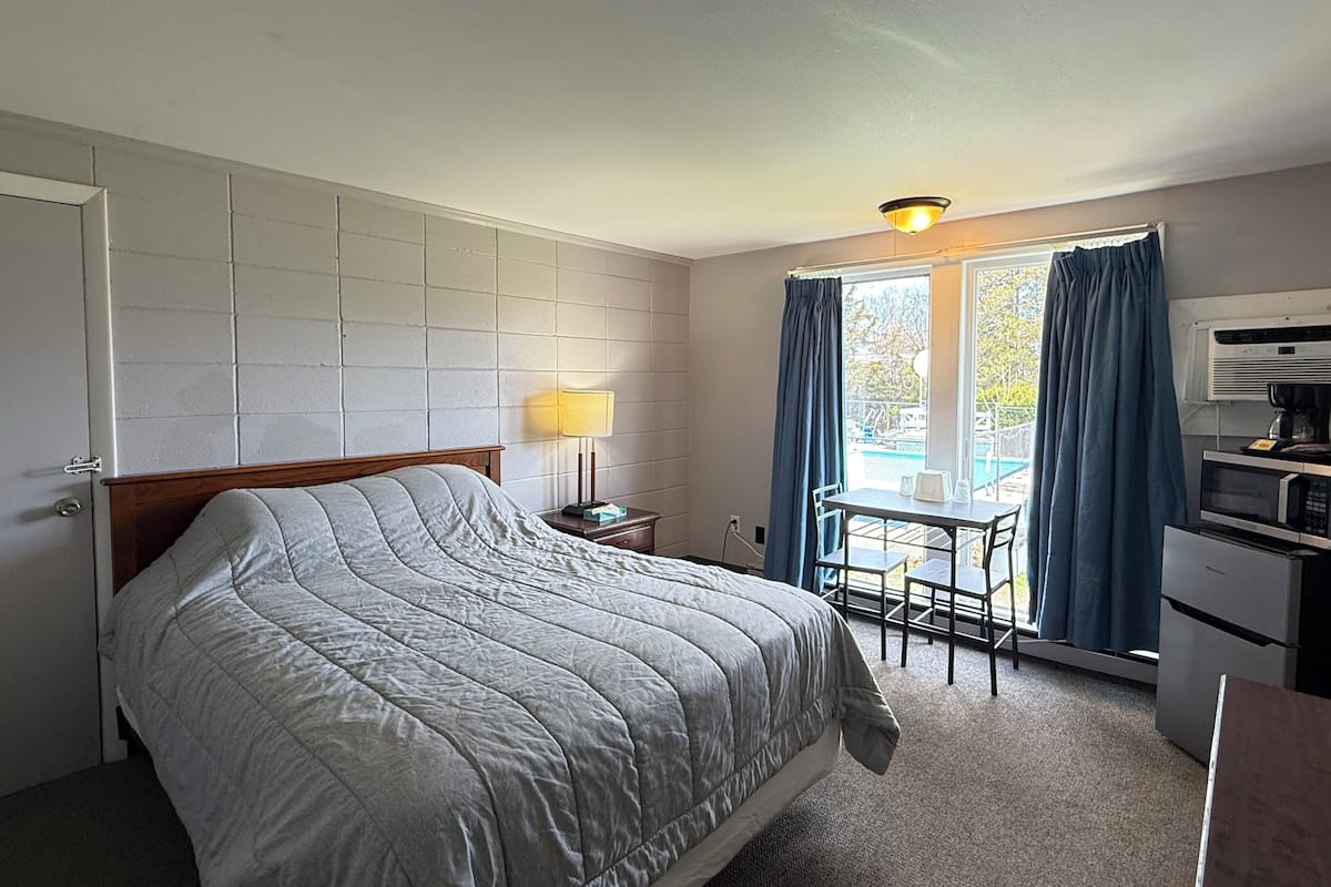 A hotel room with a bed, a desk, a chair, a lamp, a window with curtains, and a kitchenette.