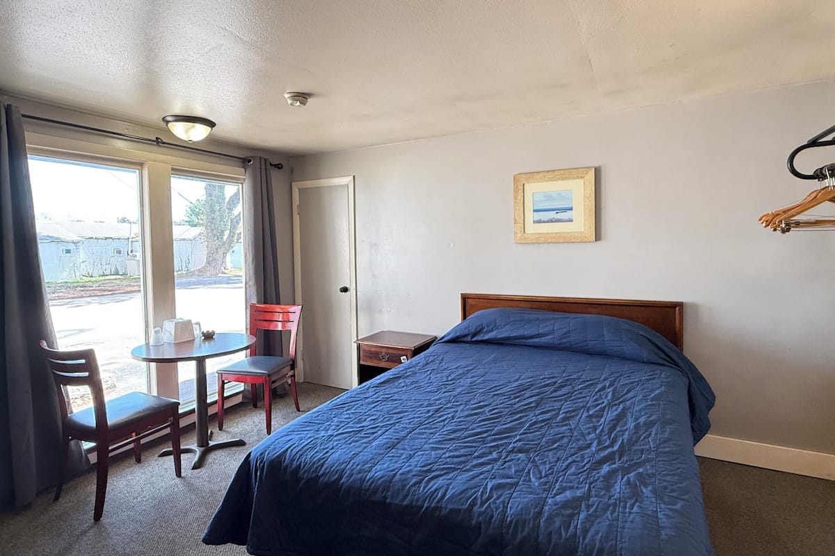 A hotel room with a bed, a small table, a chair, a coat rack, and a window with curtains.