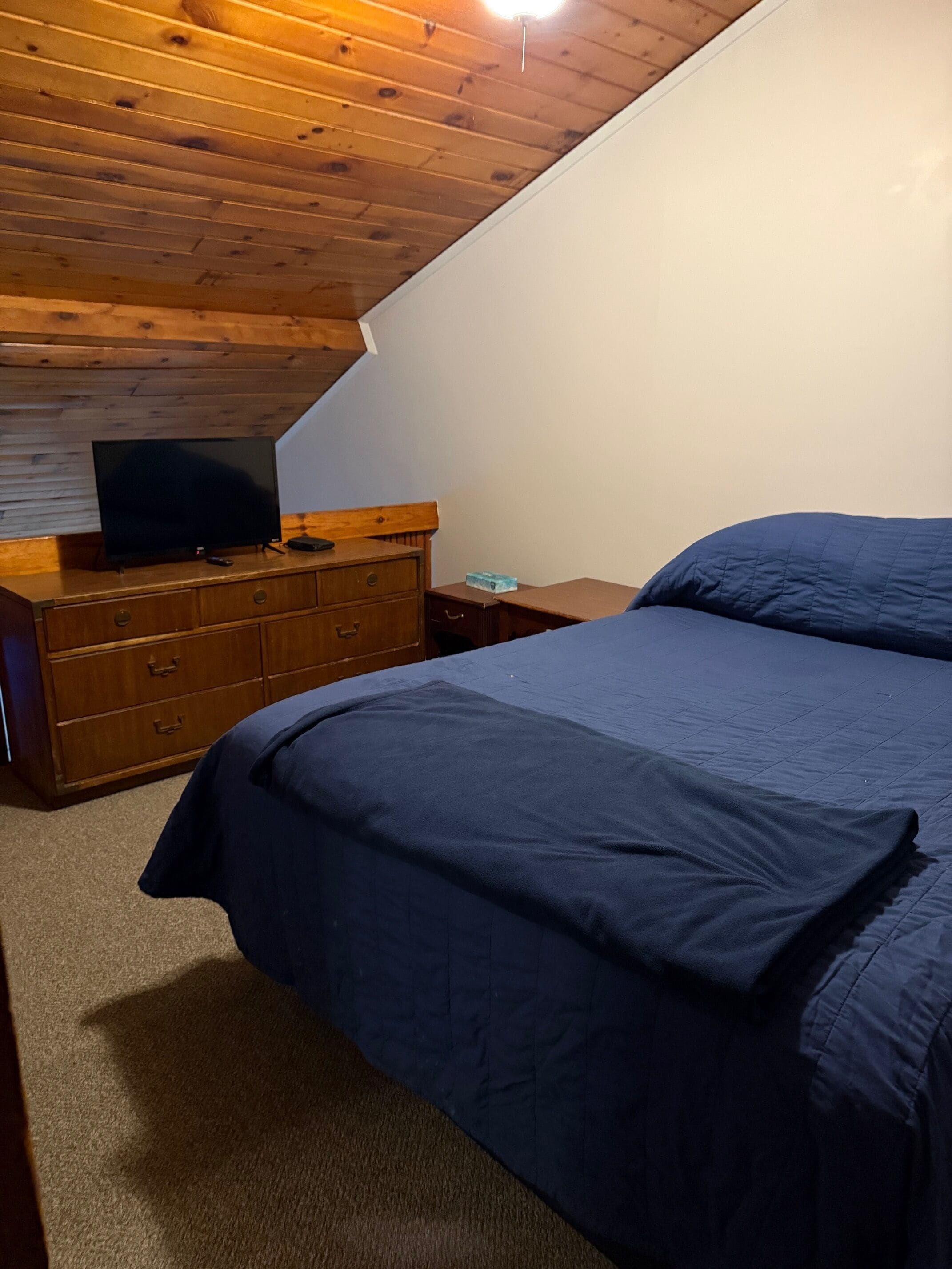Un dormitorio con techo de madera, una cama con ropa de cama azul, un televisor sobre una cómoda de madera y una mesita de noche.