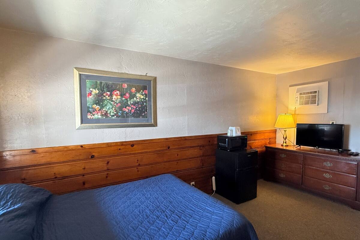 A hotel room with a bed, a wooden headboard, a desk with a lamp, a television, and a framed picture on the wall.