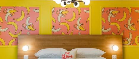 Una habitación de hotel con una cama, un almohadón con el texto 'The Grove Motel' y una pared con un estampado de plátanos, además de una lámpara en el techo.