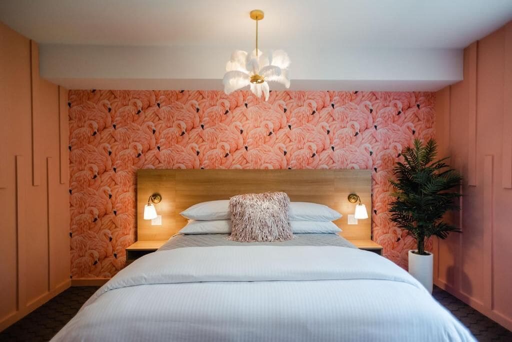 Quarto com uma cama grande, cabeceira de madeira, duas luminárias de cabeceira, parede com padrão de flamingo e uma planta em vaso.